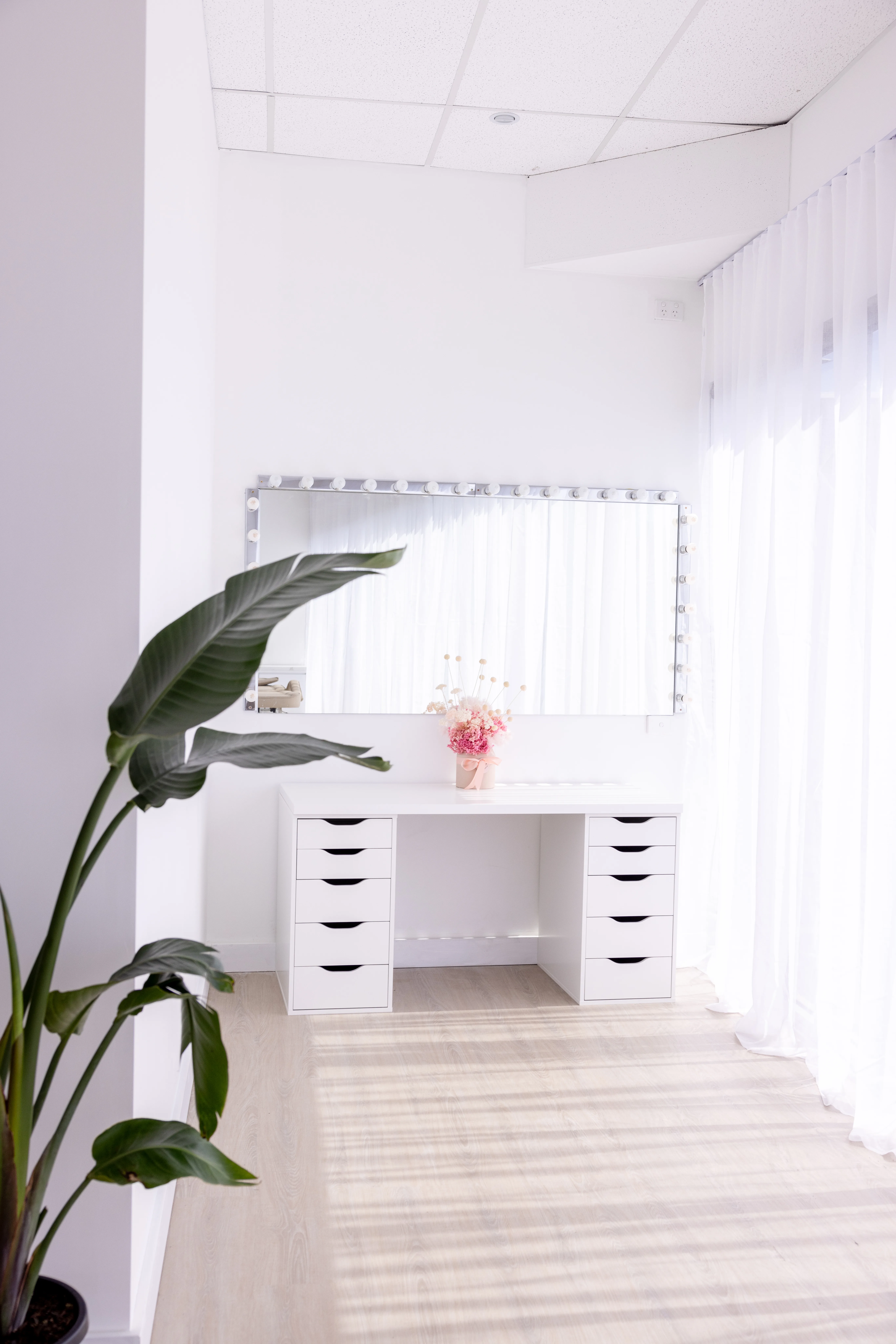 White makeup vanity with a bulb-framed mirror, dried pink florals, a bird-of-paradise plant, and sheer curtains
