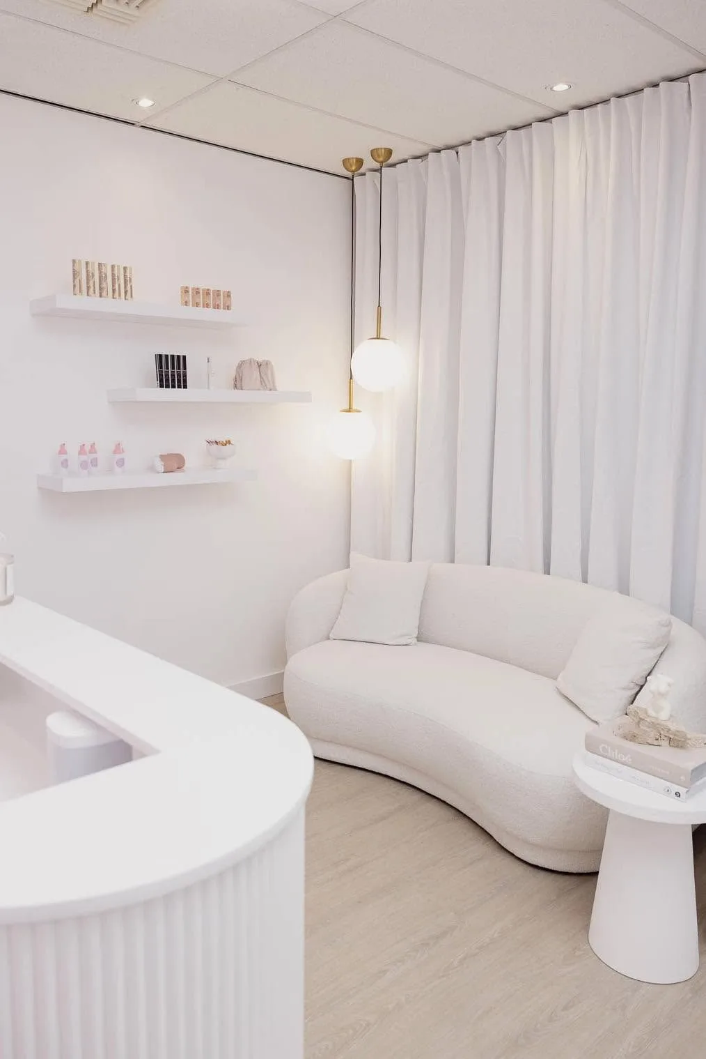 Waiting nook with a curved cream bouclé sofa, twin brass globe pendants and shelved retail product