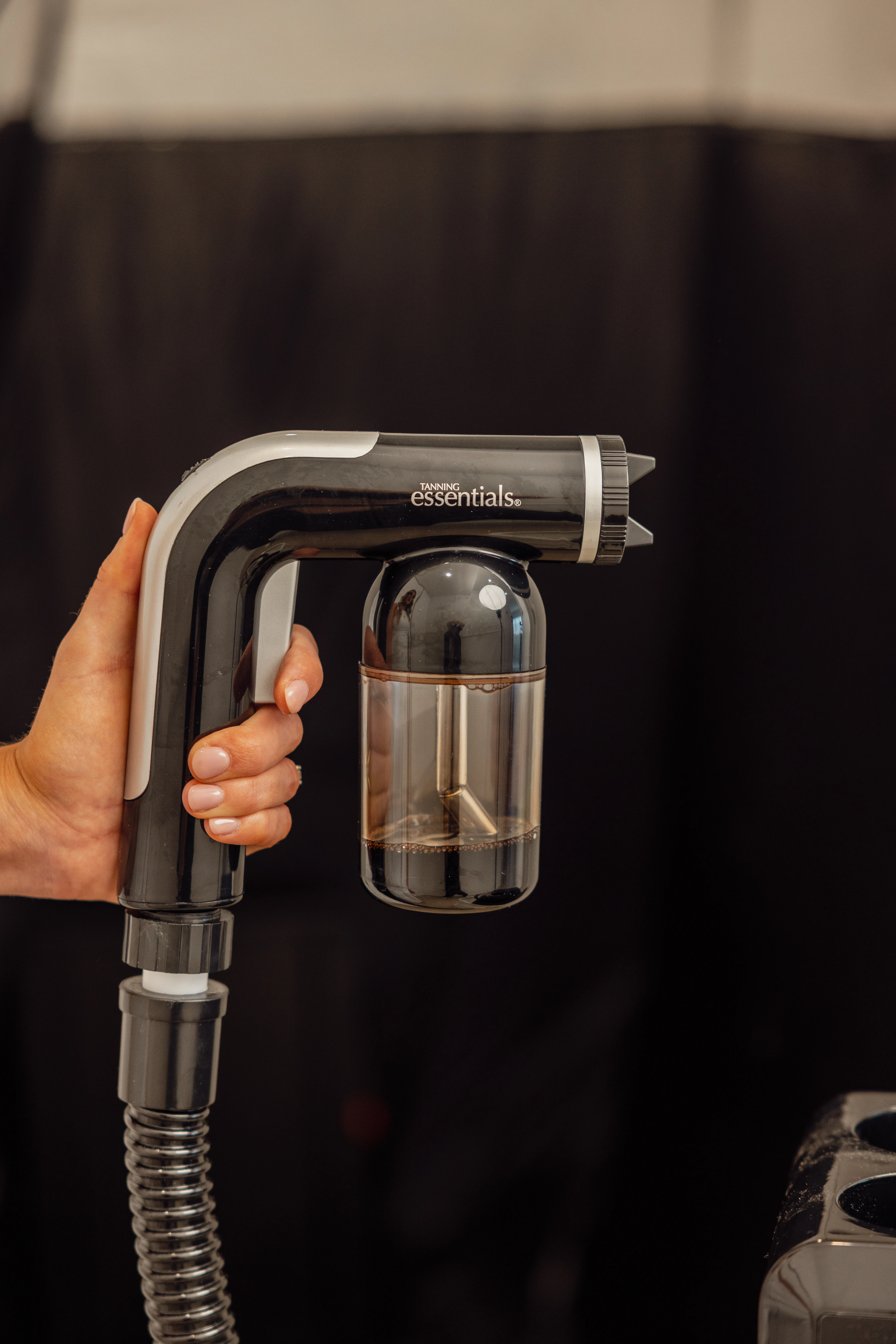 A professional Tanning Essentials spray-tan gun with a filled solution reservoir against a dark background