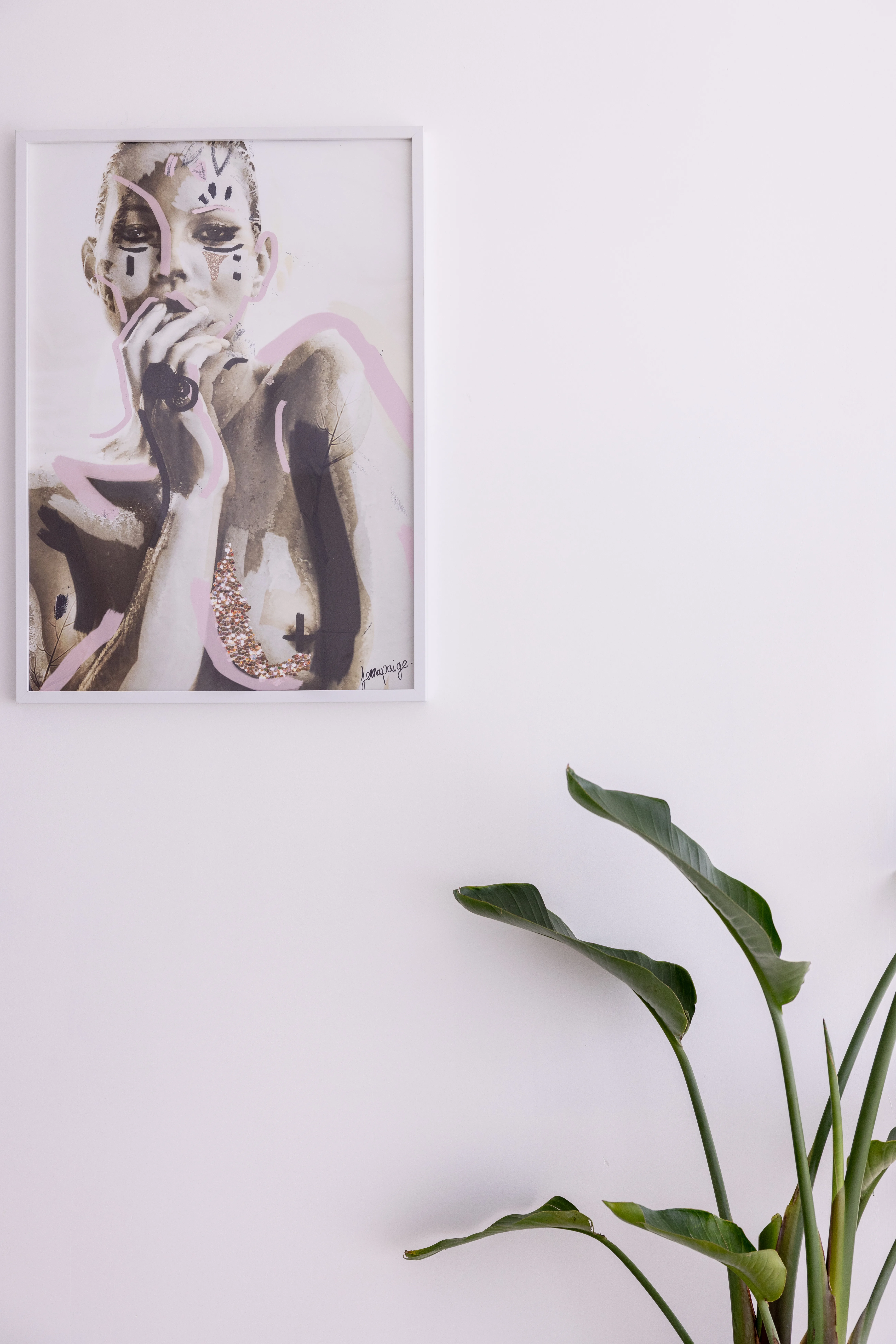 Framed fashion-illustration print of a woman in pink and gold on a blush wall, bird-of-paradise leaves entering lower right