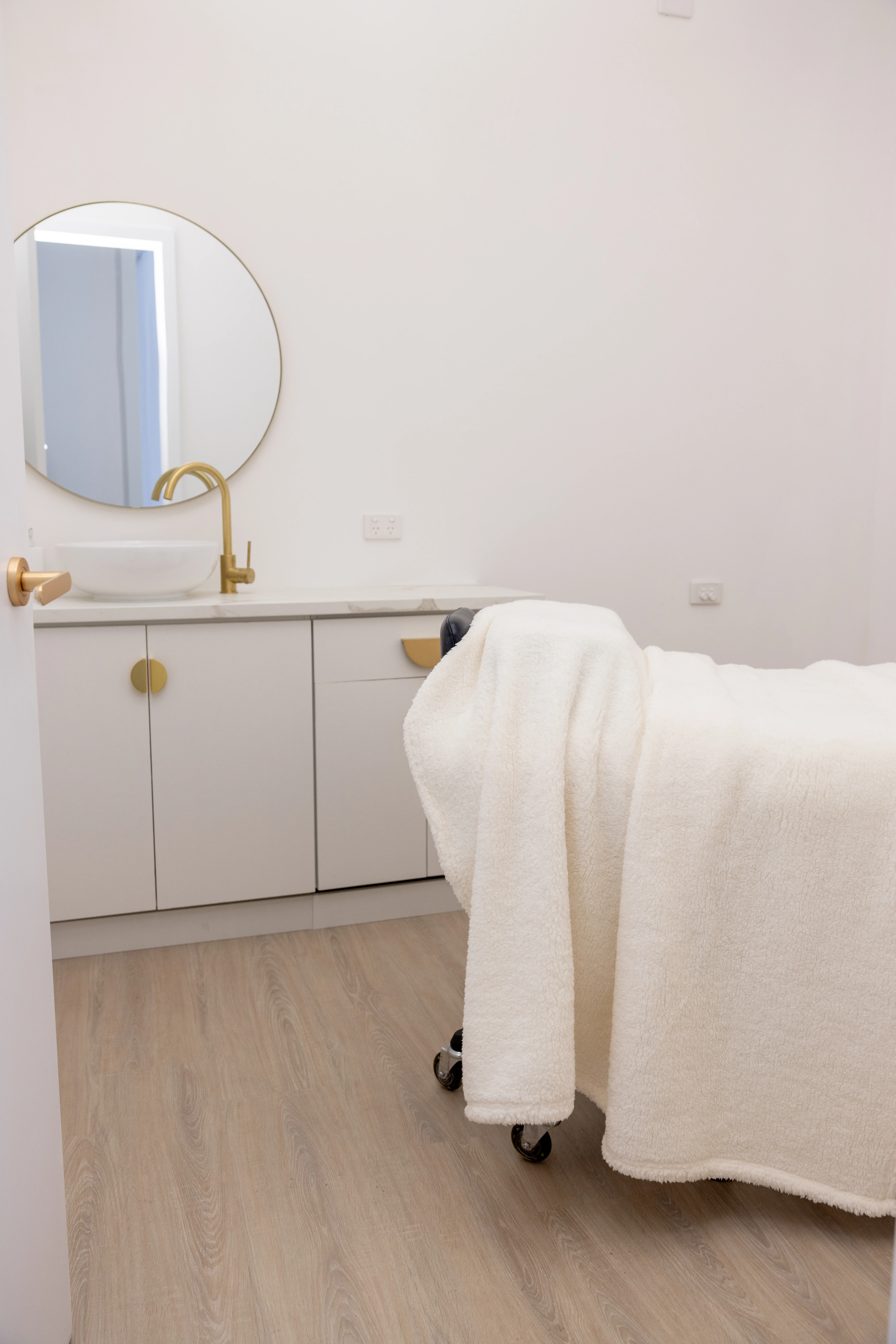 Treatment cubicle — round backlit mirror, brass tap and basin on marble, white cabinetry with brass knobs, treatment bed draped in cream towel