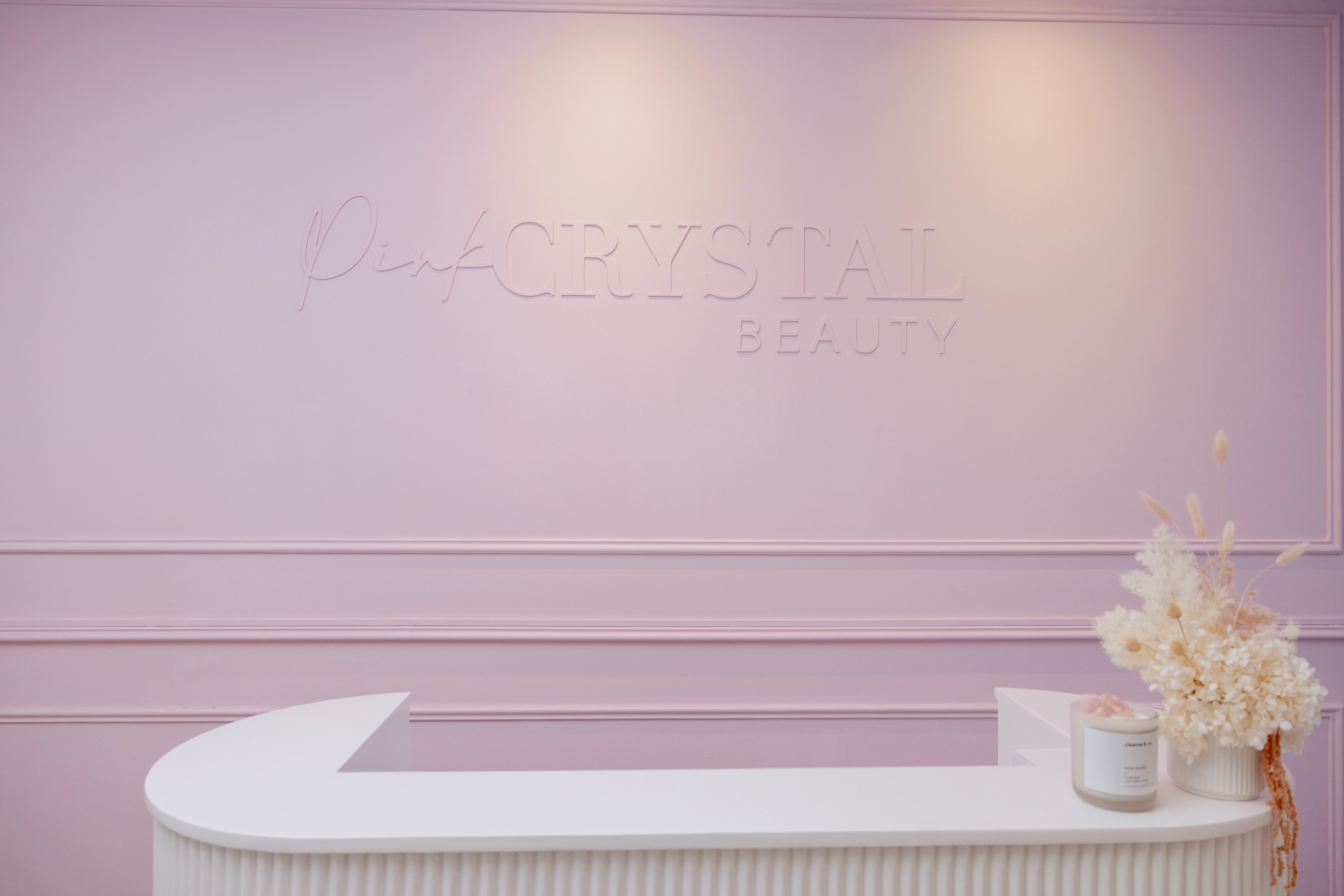 The blush wainscot wall carrying the tone-on-tone Pink Crystal Beauty wordmark above the white fluted reception desk, dried florals at right