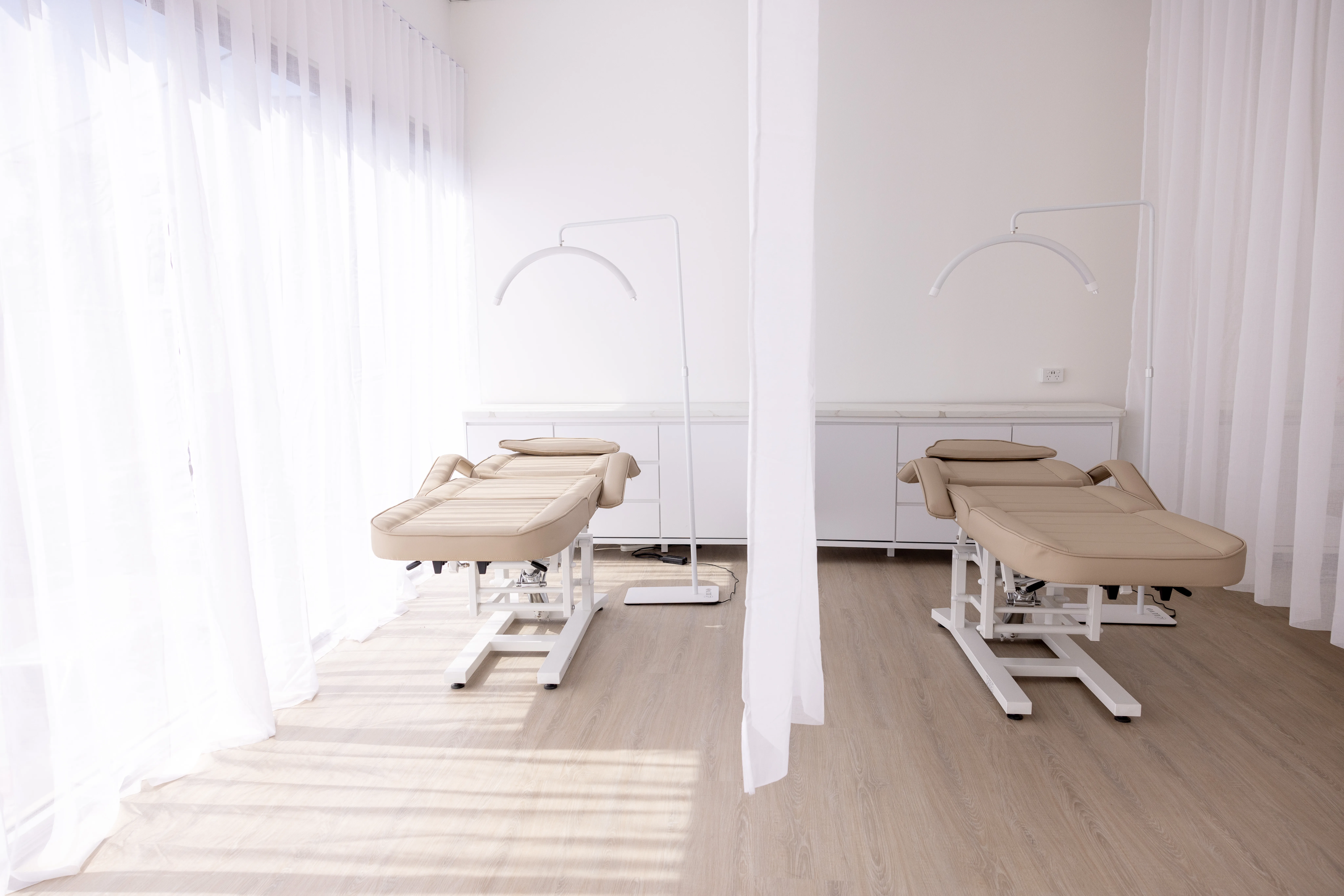 Treatment room — two beige beds under arched white LUVO lamps, sheer curtains flooding the left wall with daylight