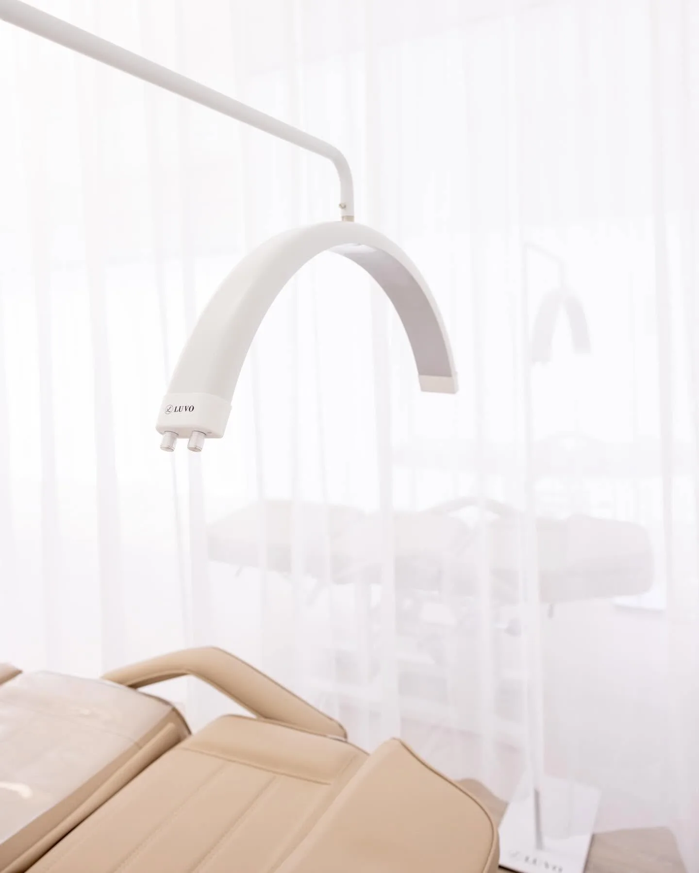 Arched white LUVO task lamp over a beige treatment bed, seen through a sheer curtain with a second bed softly veiled behind